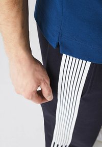 Mörkblå t-shirt med texturerad tyg och sidosprund, kombinerad med svarta byxor som har vita vertikala ränder längs sidan. Handen vilar nära byxorna.