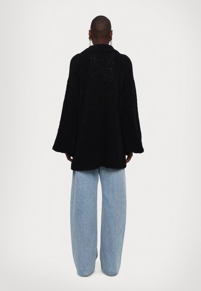 Črn strukturiran oversize kardigan z dolgimi rokavi, nošen preko svetlo modrih širokih jeans hlač. Mehka, puhasta tkanina z udobnim krojem.