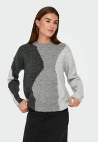 Femme portant un pull en tricot coloré en blocs de gris, noir et blanc avec des manches longues et une jupe noire, posant avec les mains près des hanches.