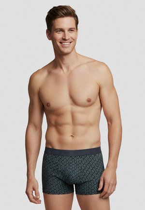 Hymyilevä, paitaton mies, jolla on vaalea iho ja ruskeat hiukset, pukeutunut tummiin kuvioituihin bokserishortseihin, seisoo yksiväristä vaaleaa taustaa vasten.