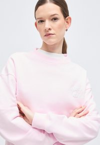 Νεαρή γυναίκα με καστανά ίσια μαλλιά σε αλογοουρά, φοράει ανοιχτό ροζ φούτερ, με σταυρωμένα χέρια, ουδέτερη έκφραση, απλό φωτεινό φόντο.