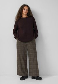 Jonge vrouw met krullend haar, gekleed in een donkerbruine trui, een bruine geblokte broek met wijde pijpen en zwarte robuuste laarzen, staande tegen een effen muur.