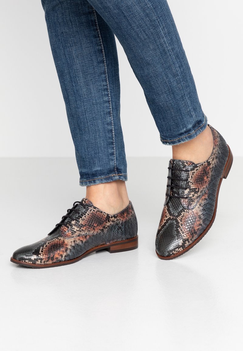 Personne portant un jean bleu et des chaussures à lacets en cuir marron-noir à motif serpent, debout sur un sol blanc.