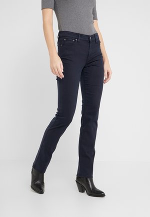 Personne portant un pantalon slim bleu foncé, un haut gris à manches courtes et des bottines noires à talons, debout devant un fond clair uni.