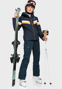 Die Skiausrüstung umfasst eine marineblaue Jacke mit orange- und weißen Streifen, passende marineblaue Hose, weiße Skischuhe und schwarze Skier mit grünen Akzenten.