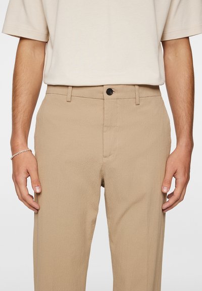 Beige Textilhose mit einem Standardbund, einer vorderen Tasche, einem einzigen schwarzen Knopf und einer lockeren Passform. Einfaches und vielseitiges Design.
