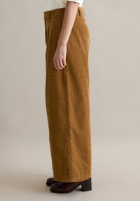 Bruine corduroy wijdvallende broek met een hoge taille, zijzakken en een vlakke voorkant; gecombineerd met zwarte blokhak schoenen.