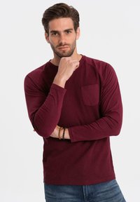 Burgundin pitkähihaisessa paidassa on ribbitekstuuri ja rintatasku, joka on yhdistetty farkkuihin. Malli poseeraa mietteliäänä käsi leualla.