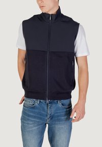 Gilet senza maniche blu navy con colletto alto, chiusura zip frontale e due tasche laterali; realizzato in un tessuto morbido e traspirante. Indossato sopra una t-shirt bianca.