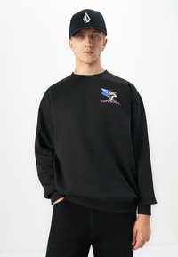 Ung man som bär en svart keps och svart sweatshirt med en färgglad tecknad grafik och "O'NEILL" text på bröstet, händerna i fickorna.