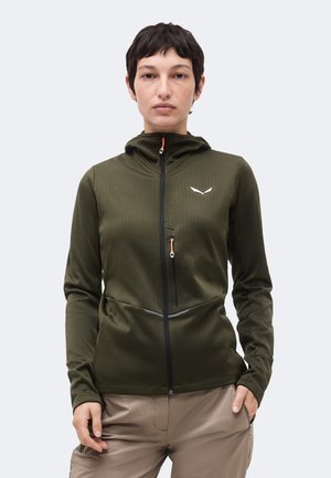 Ung person med kort mørkt hår iført en olivengrøn lynlåstrøje med en brystlomme og beige bukser, stående mod en ensfarvet baggrund.