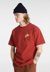 Camiseta de algodón rojo con mangas cortas y detalles en crema, con un logo de Vans amarillo en el pecho. Se lleva con un gorro navy.