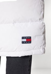 Vit puffjacka i lättviktsmaterial, med ett sydd mönster och en logopatch i svart, rött och vitt vid fållen.