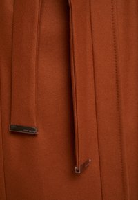 Manteau brun orange en tissu feutré doux, avec une coupe droite et sur mesure, doté de deux longues ceintures en tissu et d'accents métalliques.
