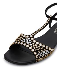 Sandal nera con molteplici dettagli in oro, caratterizzata da una suola piatta e un design aperto all'alluce, decorata con gioielli trasparenti di varie dimensioni.