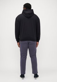 Zwarte hoodie met een zachte textuur, voorzien van een grote capuchon en een kangoeroezak, gecombineerd met grijze broek en zwarte instap schoenen.