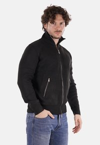 Maglioncino nero con zip e colletto alto, dotato di due tasche laterali e polsini aderenti. Il tessuto testurizzato aggiunge profondità al design.