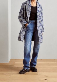 Persoon in een blauw patroonjasje, zwarte top, blauwe recht toe recht aan jeans en zwarte schoenen met een dikke zool die op een houten vloer staat.