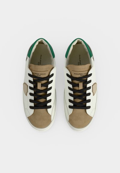 Paire de baskets blanches avec bout en daim beige, lacets noirs, détail vert au talon, vues de dessus sur un fond uni.