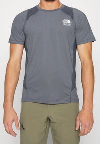 Šedé tričko s krátkým rukávem a raglánovými rukávy, s texturovanými detaily na ramenou a logem na hrudi. Hladká látka, přiléhavý design.