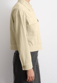 Cropped, light beige jacket in soft cotton with a pointed collar and chest pocket, featuring a button at the sleeve cuff and smooth texture.