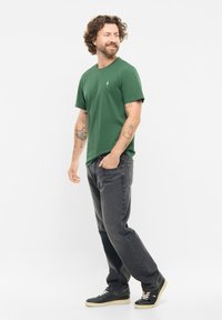 Grünes T-Shirt mit kurzen Ärmeln und kleinem Logo, kombiniert mit dunkelgrauen Jeans und schwarzen Sneakers, im lässigen Schnitt und einfarbig.