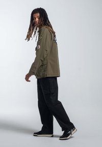 Oliwkowa kurtka wojskowa z łatami, założona na czarne spodnie cargo i czarne sneakersy z białymi podeszwami, prezentująca swobodny styl.