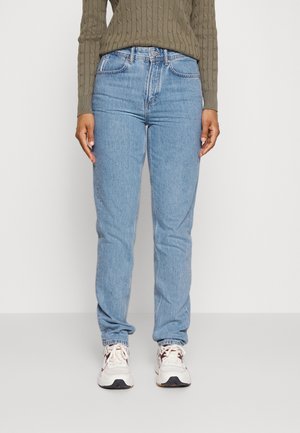 Osoba ubrana w jasnoniebieskie jeansy z wysokim stanem, oliwkowozielony sweter z warkoczowym splotem oraz białe trampki z ciemnobrązowymi detalami.