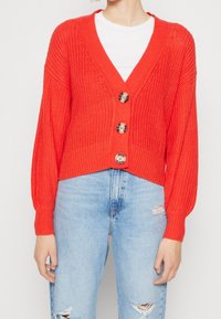 Red knitted cardigan with a V-neck, three large buttons, and long sleeves. Worn over a white shirt and paired with distressed light blue jeans.