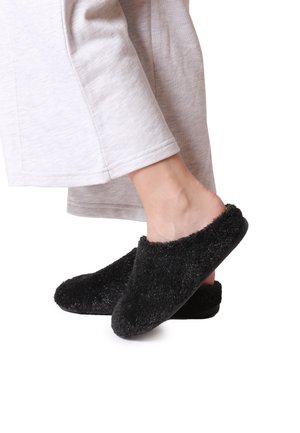 Sorte fuzzy slippers med en blød, tekstureret overflade, rund tådesign og en glat sål, båret af en person i lysegrå bukser.