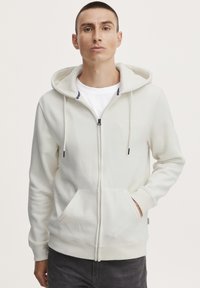 Hvit zip-up hettegenser med snor i hetten, frontlommer, marineblå detaljer på den indre kragen, og ribbede mansjetter. Myk stoffoverflate.