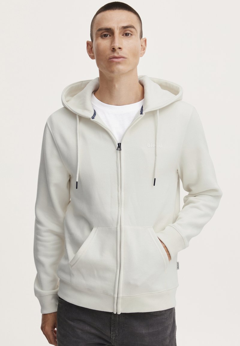 Hvit zip-up hettegenser med snor i hetten, frontlommer, marineblå detaljer på den indre kragen, og ribbede mansjetter. Myk stoffoverflate.