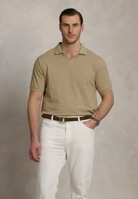 Μπεζ πόλο μπλούζα με γιακά, φορεμένη με λευκά τζιν και καφέ ζώνη. Η μπλούζα έχει κοντά μανίκια και ένα μικρό λογότυπο στο στήθος.