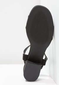 Semelle de sandale noire avec motif antidérapant texturé au talon et à l’avant-pied, montrant une bride et une boucle sur fond blanc.