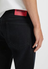 Jeans in denim scuro con una vista ravvicinata della tasca posteriore e un'etichetta in pelle rossa con la scritta "HUGO". Il design presenta un taglio classico con cuciture sottili.