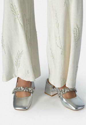 Tacones cuadrados plateados con bloque y tiras joyadas, usados con pantalones anchos color crema bordados con patrones de hojas.