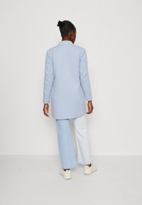 Světle modrý dlouhý kabát se střihm rovného křížení, s límcem a dlouhými rukávy, spárovaný s dvoubarevnými širokými džínami. Hladká, strukturovaná tkanina.