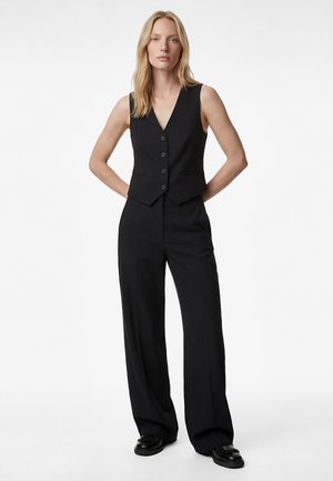 Donna con capelli biondi lunghi che indossa un gilet nero senza maniche con bottoni, pantaloni neri a gamba larga e scarpe nere, in piedi con le mani dietro la schiena.