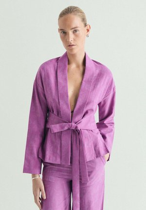 Mujer de piel clara y cabello rubio, vestida con una chaqueta morada atada y pantalones a juego, de pie frente a un fondo claro y liso.