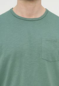 Κοντινό πλάνο ενός ατόμου που φοράει μπλουζάκι σε ανοιχτό πράσινο χρώμα με στρογγυλή λαιμόκοψη και μικρή τσέπη στο στήθος στην αριστερή πλευρά.