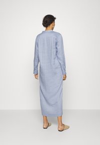 Kobieta z krótkimi, kręconymi włosami, ubrana w długą, jasnoniebieską lnianą sukienkę oraz beżowe, plecione buty wsuwane, stoi na tle jednolitego białego tła.