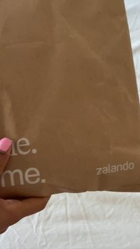 Ruda popierinė pakuotė su lygia tekstūra, ant kurios baltais rašmenimis parašyta "zalando" ir "me.", laikoma rankoje su rožiniu nagų laku.
