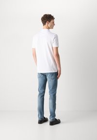 Camisa polo blanca de manga corta, con textura suave, combinada con jeans ajustados de color azul claro y zapatos negros, vista desde la parte posterior.