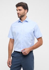 Camicia azzurra a maniche corte con bottoni in un tessuto morbido, dotata di taschino sul petto e colletto strutturato, abbinata a pantaloni scuri.
