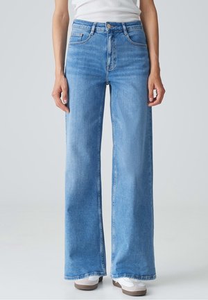 Person trägt hellblaue, weit geschnittene Jeans mit klassischem Fünf-Taschen-Design und weißen Turnschuhen, steht vor einem schlichten Hintergrund.