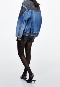 Chaqueta vaquera con paneles contrastantes en negro y azul, corte oversized, tejido texturizado, bajo elástico, combinada con pantalones cortos negros y tacones.