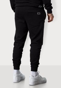 Schwarze Baumwoll-Jogginghosen mit schmalem Schnitt, ausgestattet mit zwei Taschen, elastischen Bündchen und dezentem weißen Branding am hinteren Bund und an der Tasche.