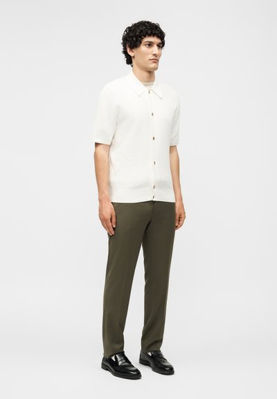 Άνδρας που στέκεται φορώντας λευκό πουκάμισο με κοντά μανίκια και κουμπιά, ελιά παντελόνια και μαύρα επίσημα παπούτσια σε απλό φόντο.