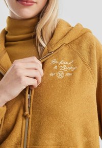 Gelbe Zip-Hoodie mit weicher Textur, versehen mit weißem, gesticktem Text "Sei nett & Glücklich" und einem Kleeblatt-Symbol auf der Brust.