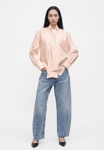 Femme debout les mains dans les poches, portant un chemisier rose à rayures avec un col noué, un jean bleu à jambes larges et des chaussures noires pointues.